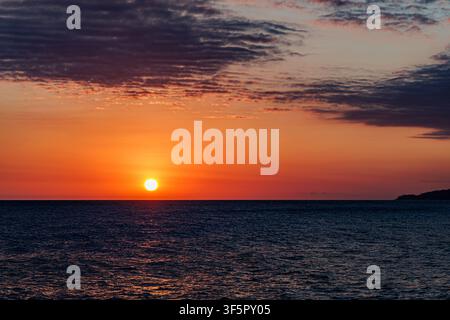 Bright golden sun setting down on horizon line over calm sea water surface under orange twilight sky. Stock Photo