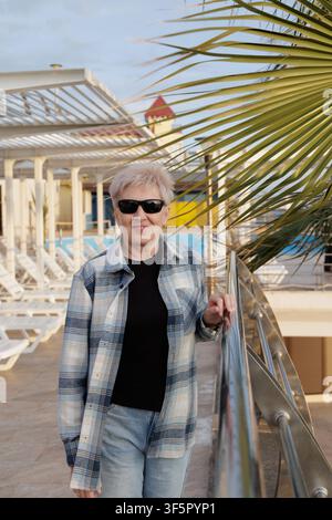 Cheerful senior female with short blonde hair in casual plaid shirt and sunglasses relaxing near palm branches at luxury hotel terrace. Stock Photo