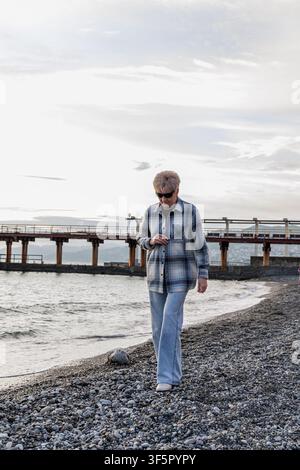 A candid medium shot of a thoughtful senior lady in a plaid jacket strolling on a coastal shore during a relaxing vacation. Stock Photo
