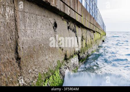 Close up of old concrete sea wall breakwater covered in green moss with ocean water wave splash at daytime. Empty copy space. Stock Photo