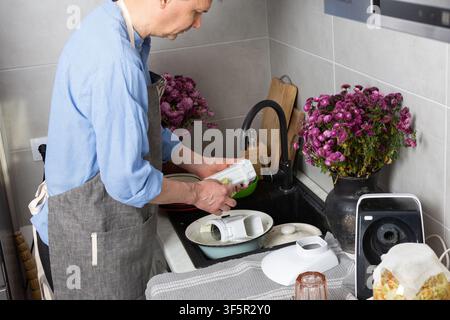 A man stands at the sink in his kitchen, cleaning dirty dishes and utensils. Flowers are placed on the counter, adding color to the scene in daylight. Stock Photo