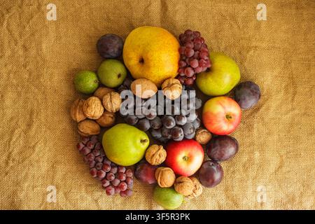 A top view of a rich autumn harvest featuring fresh organic pears, grapes, walnuts, apples, and plums arranged centrally on a rustic burlap background Stock Photo