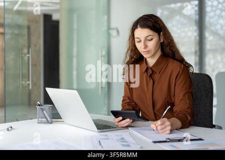 Businesswoman managing finances, analyzing data with a calculator and laptop, while taking notes on a clipboard at her modern office desk Stock Photo