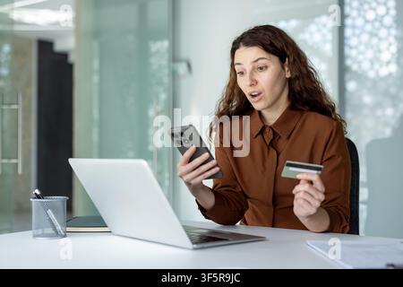 Woman reacting with surprise while holding a smartphone and credit card, using a laptop for online shopping, indicating an unexpected expense or offer Stock Photo