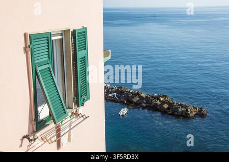 View of a window with green shutters on a pink wall overlooking the blue sea with a small boat and a breakwater in Riomaggiore, Liguria, Italy. Stock Photo