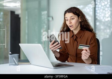 Woman reacting with surprise while holding a smartphone and credit card, using a laptop for online shopping, indicating an unexpected expense or offer Stock Photo