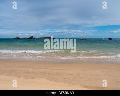 Sunlit ocean shore with gentle waves and shipwrecks on the horizon Stock Photo