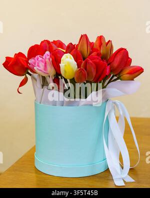 A round box holds colorful flowers with red, pink, and white blooms. The flowers sit on a wooden table in a simple room. Stock Photo