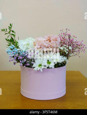 A round pink box holds a mix of flowers including hydrangeas, daisies, and gypsophila. The arrangement sits on a wooden table. Stock Photo