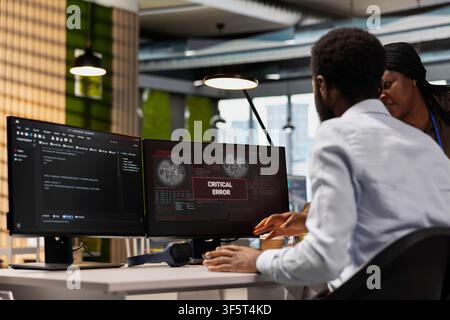 Team of system administrators working together to fix critical error warning alert in office. African american coworkers doing brainstorming, troubleshooting glitch notification on PC screen Stock Photo