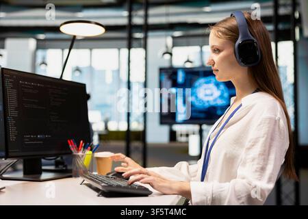 Female engineer developing applications using PC, integrating AI in office systems, listening music. Employee wearing headphones doing internship in tech startup, debugging applications Stock Photo