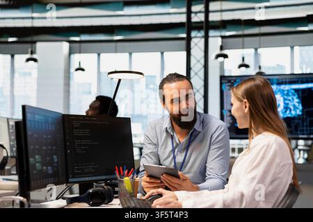 Senior engineer mentoring junior developer at workstation in office, reviewing AI system metrics. Tech company experienced manager guiding intern through code reviewing using machine learning Stock Photo
