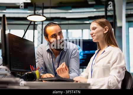 Friendly coworker having fun with young programmer while using artificial intelligence in office. Joyful colleague guiding junior dev through AI tool integration, mentoring her on using deep learning Stock Photo
