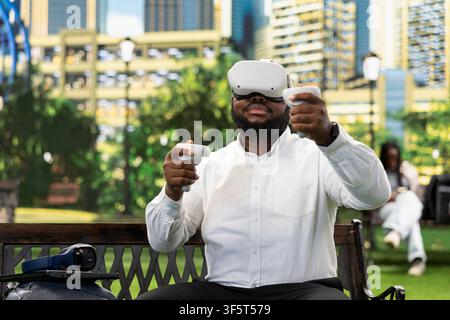 Excited businessman raises motion controller using virtual reality glasses on park bench, exploring immersive experience outdoors. Employee in public park testing VR experience. Stock Photo