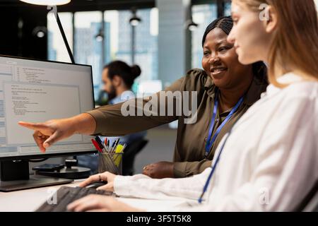 Close up of smiling IT professional helping young trainee coding during internship in tech startup. Happy senior programmer providing support to new worker using PC in software development agency Stock Photo
