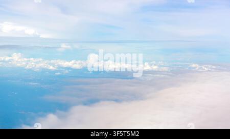 Soft layered clouds drifting above faint coastline and ocean, viewed from high altitude beneath pale blue sky with gentle sunlight, subtle haze, calm Stock Photo