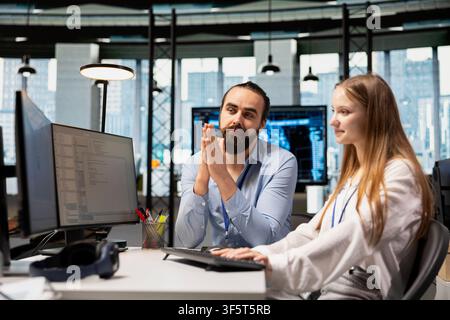 IT specialist helping young trainee coding during internship in tech startup. Lead system administrator providing support to new worker using PC in software development agency Stock Photo