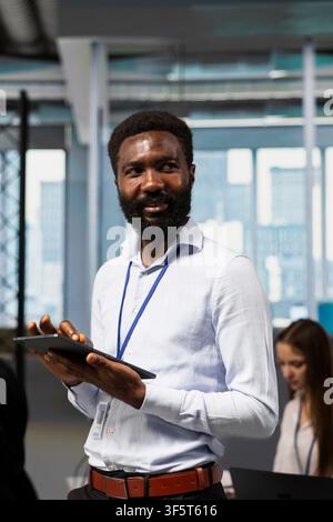 Smiling programmer using tablet to review code snippets, adjusting configurations in modern workspace. Happy employee running diagnostic tools on touchscreen device monitoring API cloud services Stock Photo