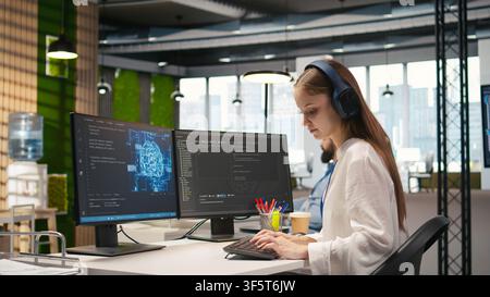 Junior dev developing software applications, optimizing code for performance, listening music. Young female engineer wearing headphones in office using AI to resolve bottlenecks, camera A Stock Photo