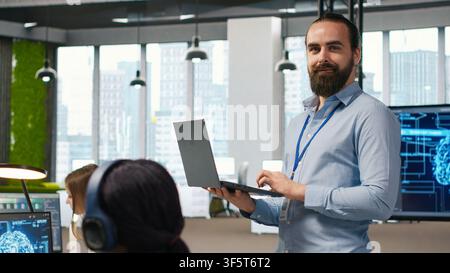 Jolly programmer coding AI algorithms on laptop to debug software issues and improving system stability. Smiling employee in tech startup office configuring settings using notebook, camera A Stock Photo