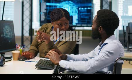 Cheerful manager and employee with paraplegia in inclusive workplace developing software applications. African american technician and colleague with disability using AI to solve bottlenecks, camera A Stock Photo