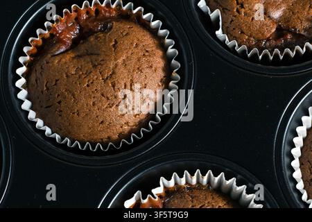 Chocolate muffins rest in a dark tray with white paper cups. Brown tops show cracked texture and risen batter. Closeup view supports food content. Stock Photo