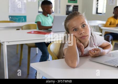 Diverse elementary students sitting at classroom desks using laptops with red notebook and posters. School, children, computers, tables, chairs, bright, windows Stock Photo