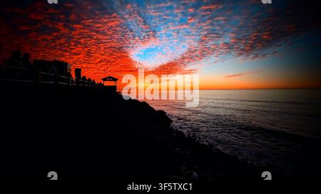 Dramatic fiery sunset over the ocean horizon featuring vibrant red altocumulus clouds and silhouetted people watching from coastal promenade pavilion Stock Photo