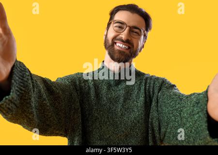 Portrait of happy young man in warm sweater taking selfie on yellow background Stock Photo