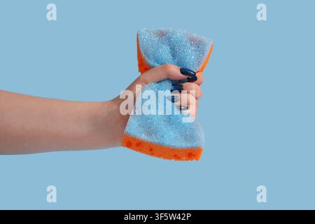 Female hand squeezing a blue and orange cleaning sponge with dark blue glitter nail polish on a light blue background, the hand has dark blue glitter Stock Photo