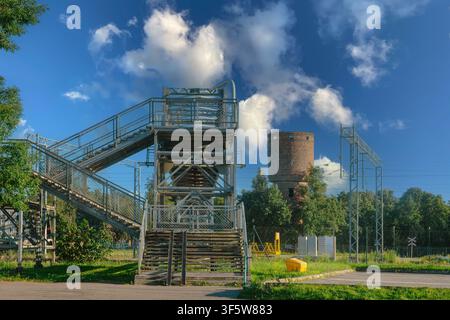 Steel pedestrian overpass with wooden steps and metal railings crossing railway area brick industrial tower emitting steam in background green trees p Stock Photo