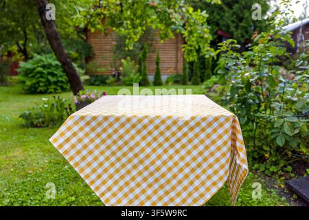 Empty table covered with a white and yellow checkered cotton tablecloth in a lush green summer garden. Bright outdoor scene with natural light and gen Stock Photo