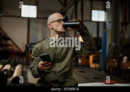 Male Welder Drinking Water and Using Smartphone during Work Break Stock Photo