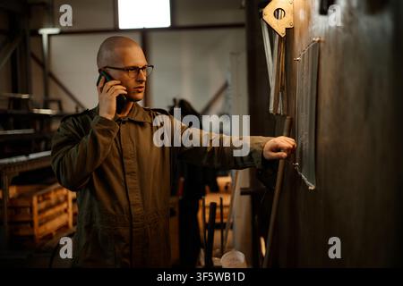 Male Welder Talking on Smartphone and Examining Blueprint in Workshop Stock Photo