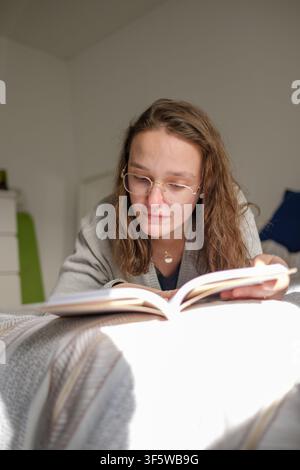 Young woman reading a book with glasses lying on the bed during leisure time Stock Photo
