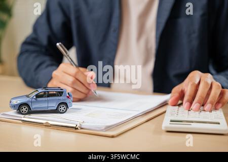A man signs car insurance documents, calculating costs with a loan calculator. Writing his signature on a contract, ensuring smooth financial transact Stock Photo