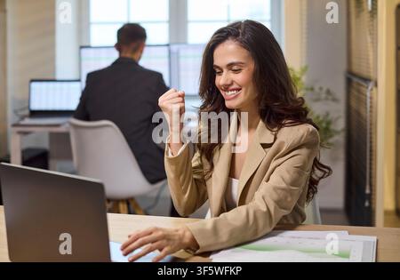 Joyful businesswoman celebrating sales success at laptop in bright modern office Stock Photo