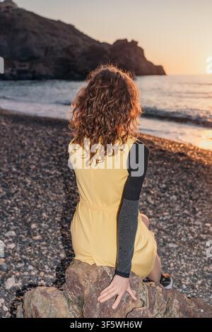 Woman sunset beach sits on a coastal rock watching waves during golden hour on a peaceful pebble shore Stock Photo