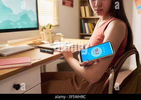 Girl Scanning Glucose Sensor on Arm with Smartphone at Desk Stock Photo