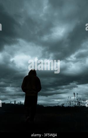 Anonymous hooded figure walking alone beneath heavy storm clouds in a dark rural landscape, seen from behind Stock Photo