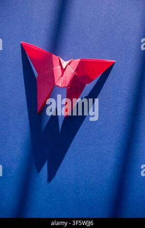 Red origami butterfly casting a long geometric shadow on a deep blue background in strong directional light. Stock Photo