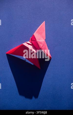 Red origami butterfly casting a long geometric shadow on a deep blue background in strong directional light. Stock Photo