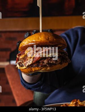 Hand holding a double smash beef burger with melted cheese, smoked beef bacon and burger sauce in a glazed brioche bun, in a dimly lit restaurant. Stock Photo