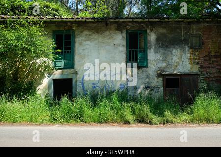 Abandoned rustic country house with weathered green wooden doors and graffiti on the wall by a rural road in Asturias, Northern Spain. Stock Photo