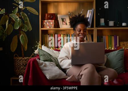 Smiling young adult African American woman using laptop on sofa at home, working remotely or studying online. Image suitable for remote work, e-learning, and digital lifestyle marketing Stock Photo