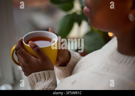 African American young woman holding mug of hot tea near window, practicing mindfulness and self care. Image suitable for mental wellbeing campaigns and lifestyle marketing, close up shot Stock Photo