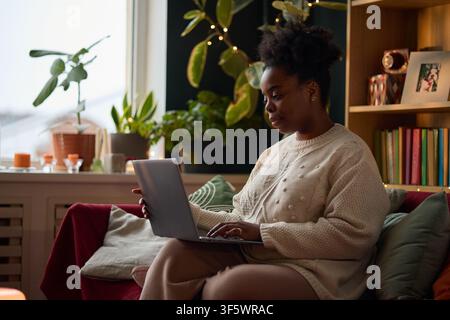 African American young adult woman sitting on sofa and typing on laptop, working from home. Image represents remote work, online education, and e-learning, suitable for corporate marketing Stock Photo