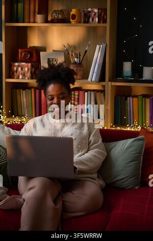Vertical image of young adult African American woman smiling while using laptop on sofa at home. Concept of remote work, online education, or e-commerce shopping Stock Photo