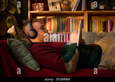 Young adult Caucasian man wearing headphones lying on sofa and writing in notebook, studying at home. Image represents remote learning and mental wellbeing, ideal for education marketing Stock Photo