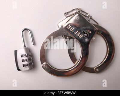 An open silver combination padlock placed next to shiny metal handcuffs on a plain white surface. Stock Photo